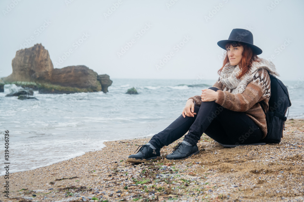 Beautiful red-haired woman in a hat and scarf with a backpack sits on the coast against the background of the rocks against the beautiful sea. Tourism, rest, lifestyle. Place for text or advertising