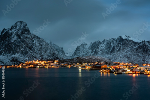 Reine village harbor at night with building's lights and outstanding snowy mountains. Lofoten islands, Norway.
