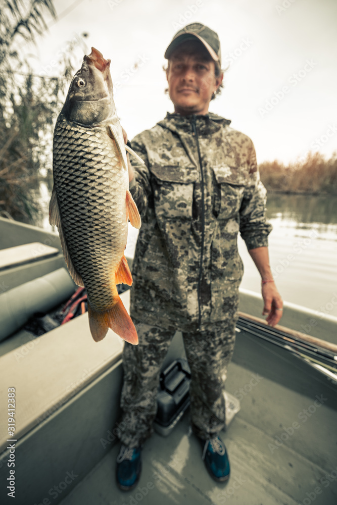Naklejka premium Young amateur angler holds the big Carp fish (Cyprinus carpio) being in the boat on the river