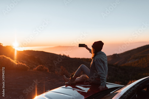 Young woman photographing with phone beautiful landscape during a sunset, sitting on the car hood while traveling high in the mountains