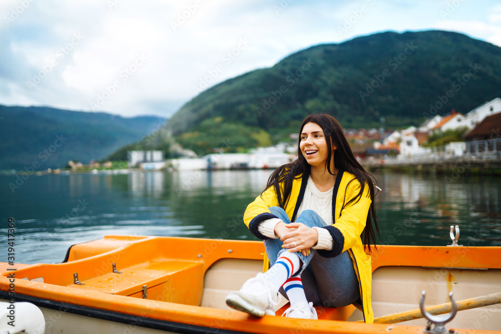 The girl tourist in a yellow jacket is sitting and posing in a boat against the backdrop of the mountains in the Norway. Young woman relaxing on the boat by the lake. Travelling, lifestyle, adventure. The girl tourist in a yellow jacket is sitting and posing in a boat against the backdrop of the mountains in the Norway. Young woman relaxing on the boat by the lake. Travelling, lifestyle, adventure.