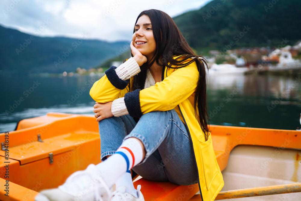 The girl tourist in a yellow jacket is sitting and posing in a boat against the backdrop of the mountains in the Norway. Young woman relaxing on the boat by the lake. Travelling, lifestyle, adventure.