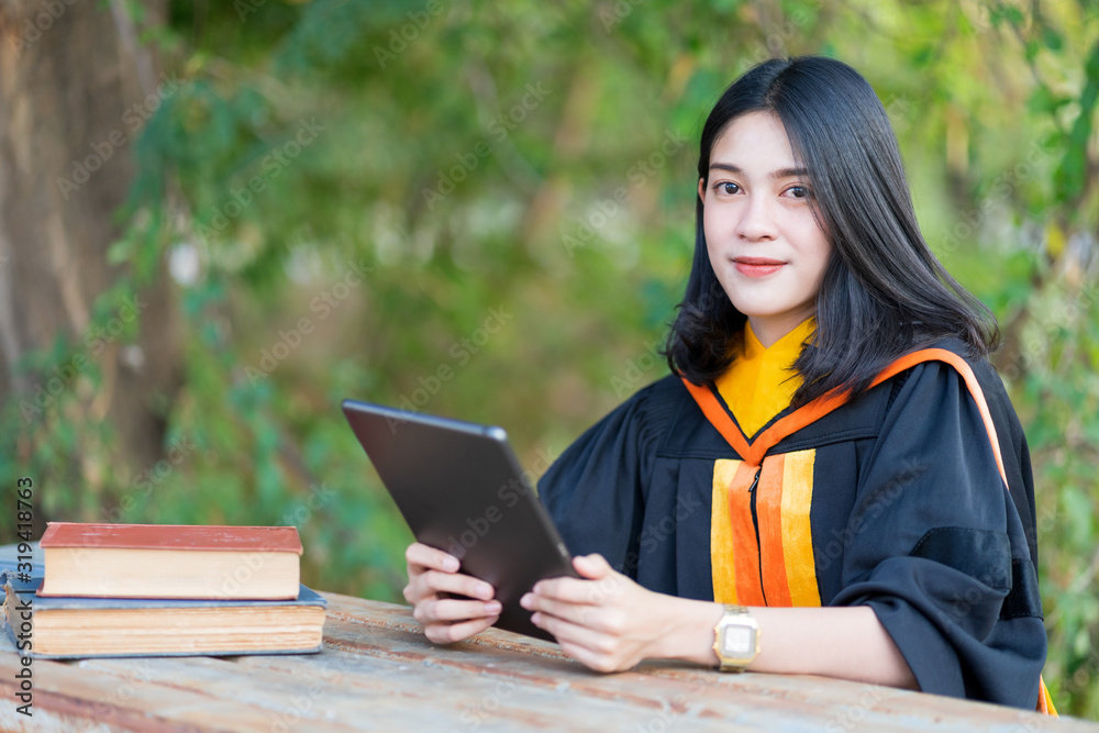 Fototapeta premium A young cheerful beautiful female gradute student wearing academic gown studying in college campus with books and lablet on table.