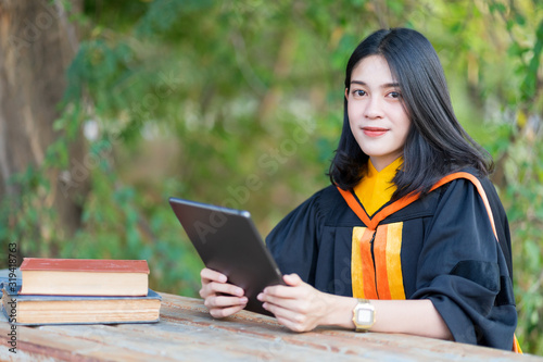 A young cheerful beautiful female gradute student wearing academic gown studying in college campus with books and lablet on table.
