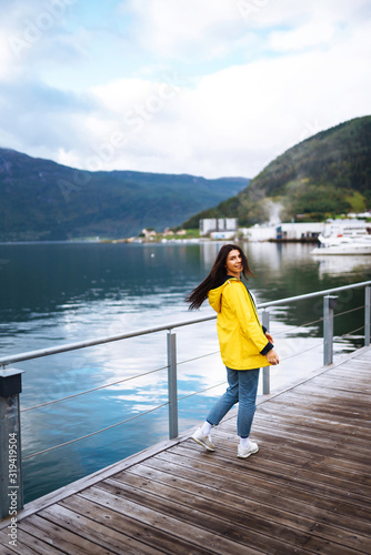 The girl tourist in a yellow jacket walking along the promenade in the Norway. Young woman smilling and posing against the backdrop of the mountains. Travelling, lifestyle, adventure concept.