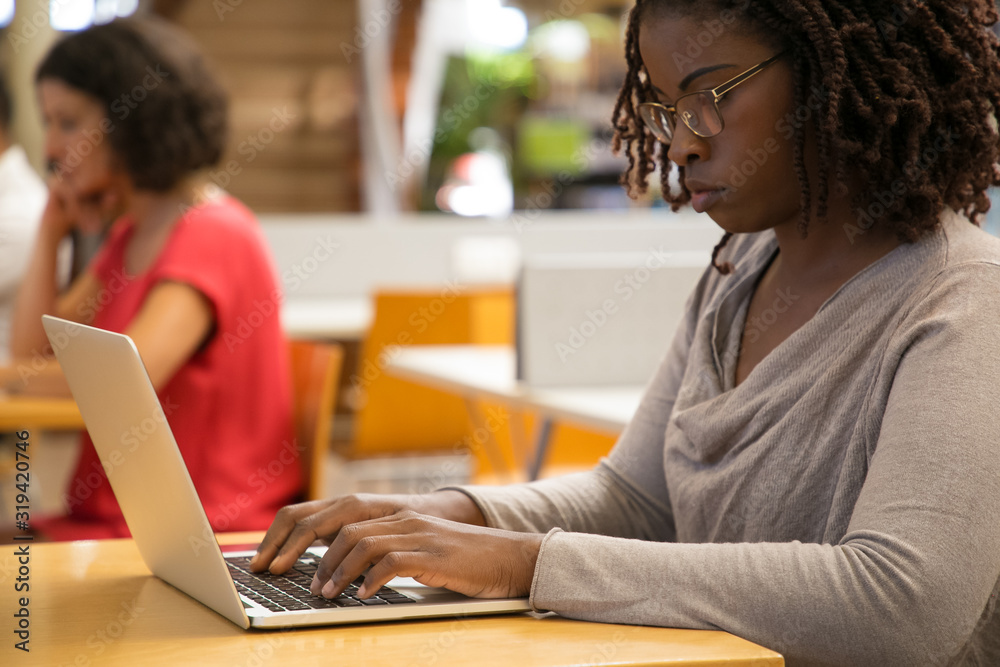 Fototapeta premium Thoughtful woman working with laptop at public library. Concentrated African American woman typing on laptop. Technology concept