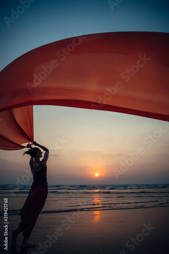 Elegant young girl performs dance moves on the sand holding a long red cloth against the background of evening sky and sun setting over the sea. Silhouette of slim woman against the sunset.