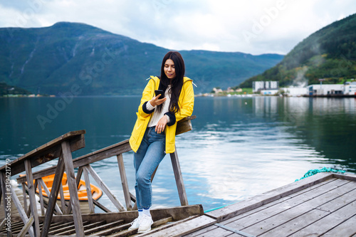 The girl tourist takes a photo on the phone by the lake in Norway. Young woman takes selfie against the backdrop of the mountains in the Norway. Travelling, lifestyle, adventure, concept. © maxbelchenko
