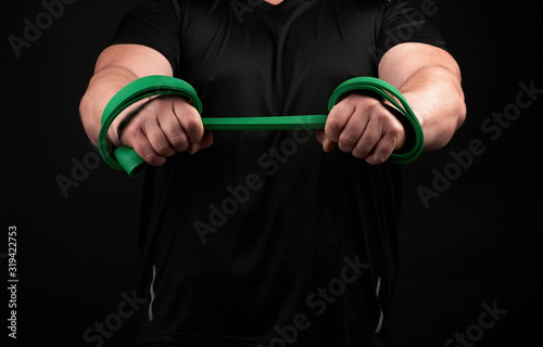 athlete with a muscular body in black clothes is doing physical exercises with green rubber