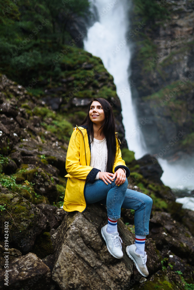 The girl tourist in a yellow jacket enjoys freedom against the backdrop of the waterfall. Young woman looking on the majestic waterfall in the Norway. Travelling, lifestyle, adventure, concept.