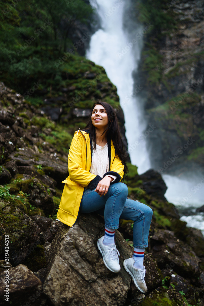 The girl tourist in a yellow jacket enjoys freedom against the backdrop of the waterfall. Young woman looking on the majestic waterfall in the Norway. Travelling, lifestyle, adventure, concept.