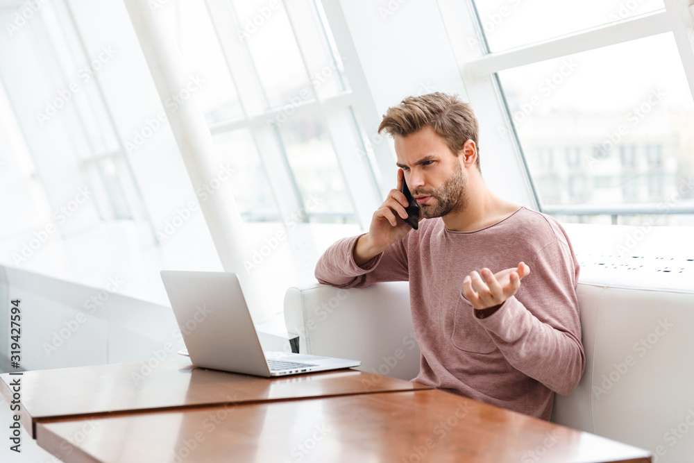 Image of young bearded man using smartphone and laptop in cafe indoors Image of young bearded man using smartphone and laptop in cafe indoors