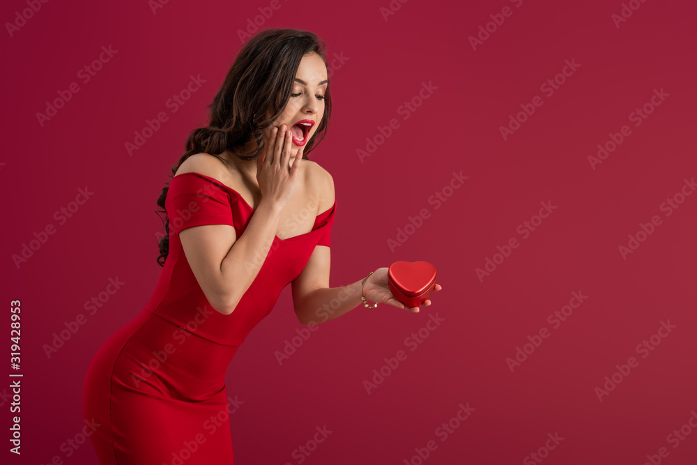 surprised, elegant girl touching face while holding heart-shaped gift box isolated on red surprised, elegant girl touching face while holding heart-shaped gift box isolated on red