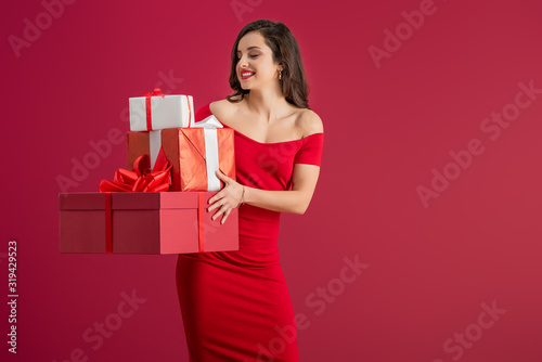 sexy, elegant girl holding gift boxes while smiling isolated on red