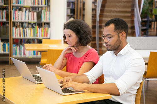 Two concentrated students using laptops while sitting at library. Focused people studying with laptops together. Technology, education concept