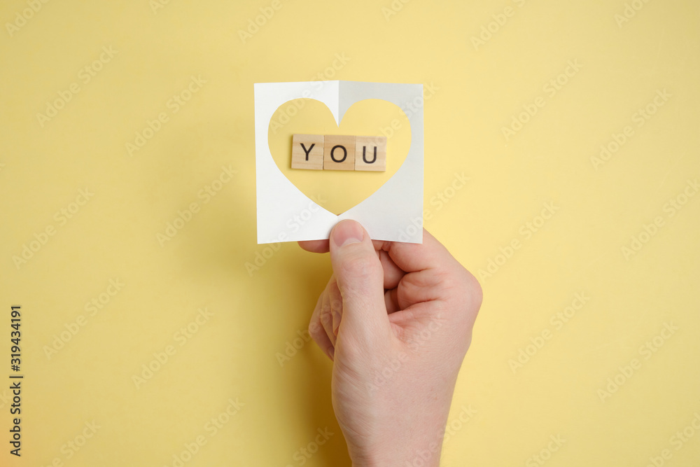 Declaration of love. Hand holds abstract paper heart over wooden squares with the word - you. Paper yellow background. Declaration of love. Hand holds abstract paper heart over wooden squares with the word - you. Paper yellow background.