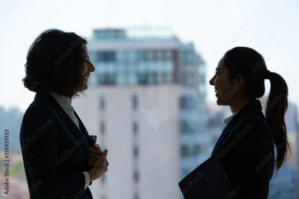 Happy businesswomen talking in office. Side view of cheerful multiethnic female colleagues standing together and talking in modern office. Cooperation concept