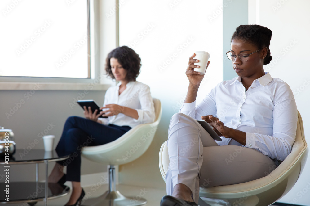 Businesswomen drinking coffee and using digital tablets. Multiethnic female colleagues sitting in chairs and using tablet computers in office during coffee break. Technology concept Businesswomen drinking coffee and using digital tablets. Multiethnic female colleagues sitting in chairs and using tablet computers in office during coffee break. Technology concept