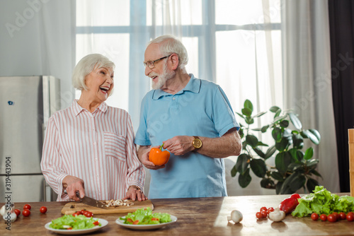 Selective focus of senior woman laughing while cutting vegetables by husband in kitchen