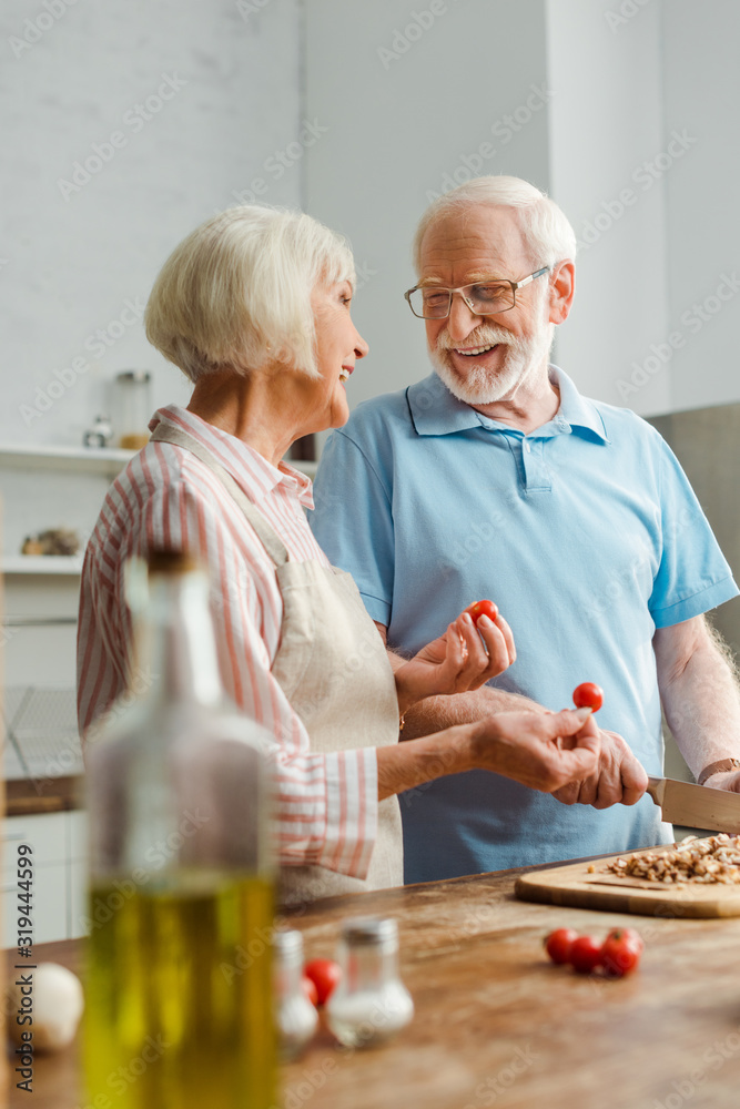 Selective focus of smiling man looking at wife with cherry tomatoes while cutting mushrooms on kitchen table