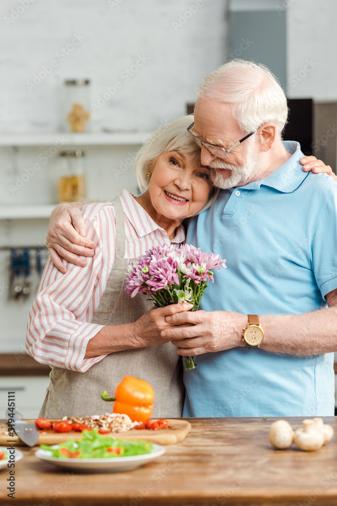 Senior woman with bouquet hugging husband and looking at camera beside fresh vegetables on kitchen table Senior woman with bouquet hugging husband and looking at camera beside fresh vegetables on kitchen table