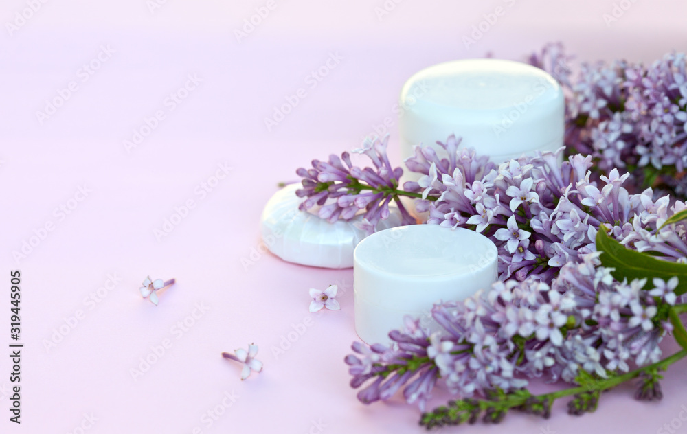 Two jars of moisturizer for face and body care and a cosmetic soap with sprigs of blooming lilacs on a pink background. Spring romantic still life. Close-up, blanc, mock up, macro, place for text Two jars of moisturizer for face and body care and a cosmetic soap with sprigs of blooming lilacs on a pink background. Spring romantic still life. Close-up, blanc, mock up, macro, place for text