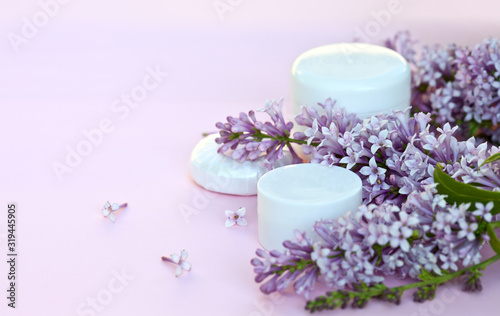 Two jars of moisturizer for face and body care and a cosmetic soap with sprigs of blooming lilacs on a pink background. Spring romantic still life. Close-up  blanc  mock up  macro  place for text
