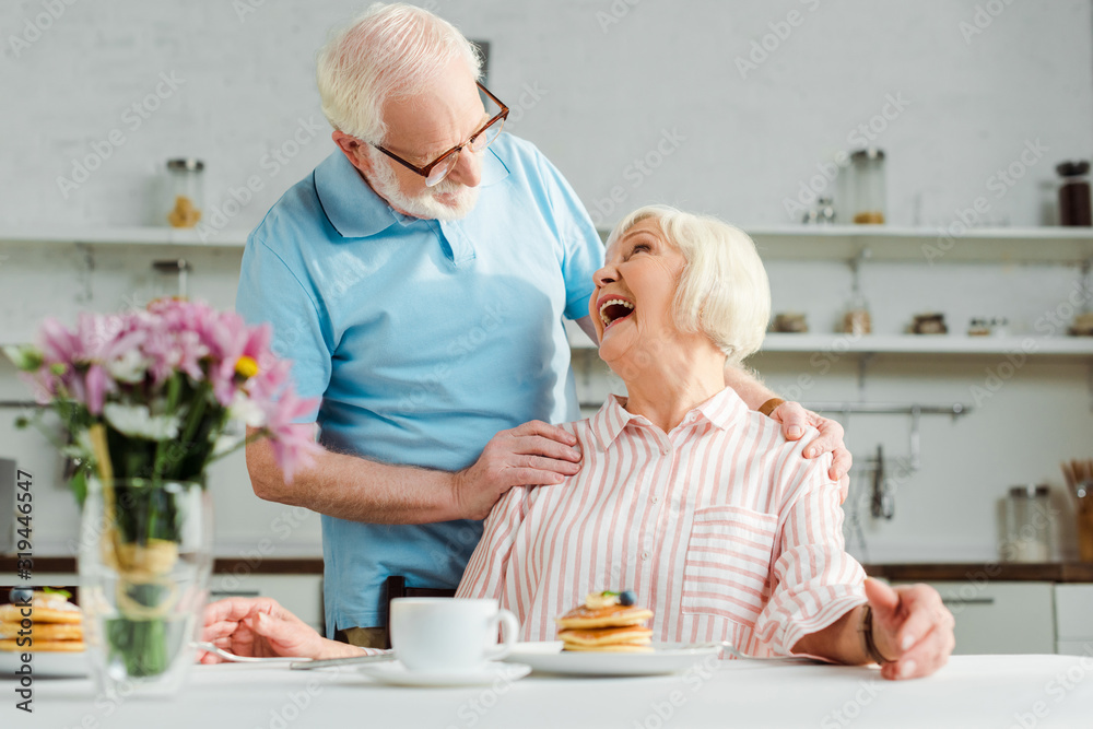 Selective focus of senior woman laughing while looking at husband by coffee and pancakes on table in kitchen