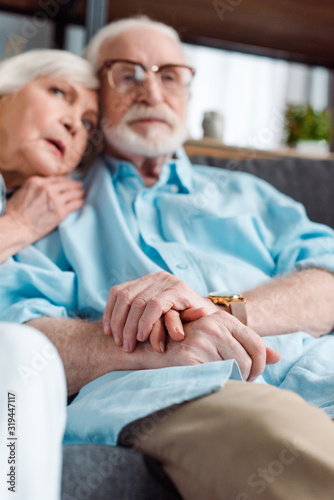 Selective focus of senior couple holding hands while sitting on sofa at home