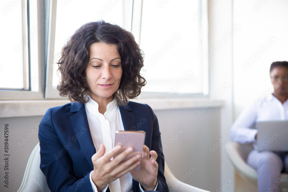 Focused middle aged businesswoman using smartphone. Selective focus of businesswoman using mobile phone while female colleague working with laptop in modern office. Business and technology concept Focused middle aged businesswoman using smartphone. Selective focus of businesswoman using mobile phone while female colleague working with laptop in modern office. Business and technology concept