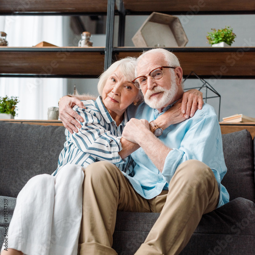 Senior couple looking at camera while hugging on couch