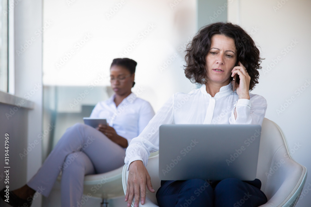 Serious businesswoman using laptop and talking by smartphone. Businesswoman working with laptop computer and cell phone while female colleague sitting behind in office. Business and technology concept Serious businesswoman using laptop and talking by smartphone. Businesswoman working with laptop computer and cell phone while female colleague sitting behind in office. Business and technology concept