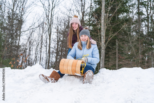 Fotografie Two happy teen girls/friends sitting on a wood toboggan/sled while at the edge of a snow covered hill in a park