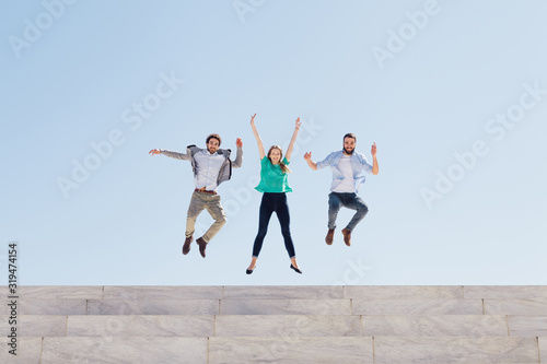 Three friends jump into the air at the top of an external staircase of a building, behind them the blue sky of a spring day - Copy space - Millennial have fun together