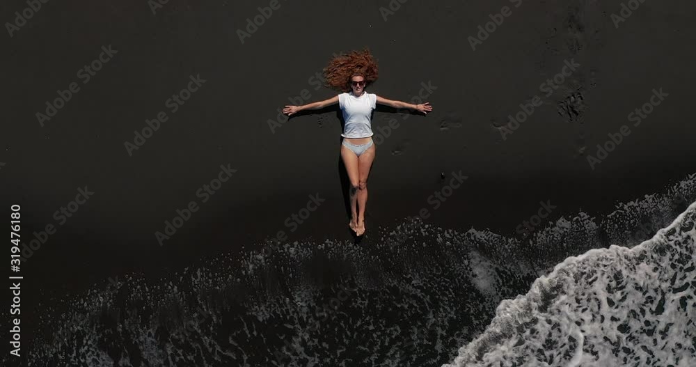 Aerial view. Beautiful woman lay down and rest on the black beach in tenerife while a long oceanic wave is coming.
