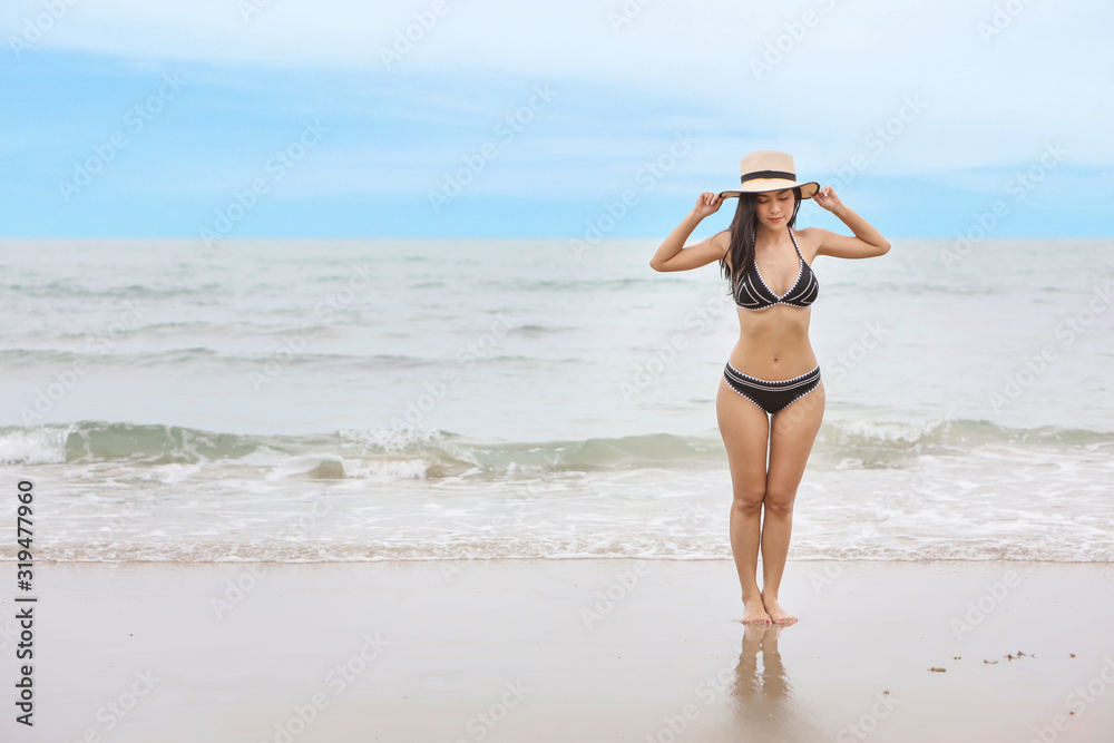 Portrait of young asian beautiful and sexy woman in black bikini standing on the beach and holding hat. Cute girl having fun on holiday vacation with happy smiling face. Free space for advertising. . Portrait of young asian beautiful and sexy woman in black bikini standing on the beach and holding hat. Cute girl having fun on holiday vacation with happy smiling face. Free space for advertising. .