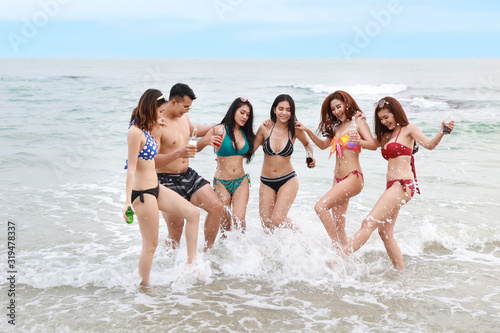 Group of happy young asian women and man wearing bikini or swimming suit. They kicking water, holding alcohol and soft drink bottles on sandy beach party together and having fun. Friendship concept
