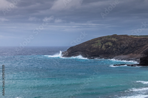 Oahu, Hawaii, USA. - January 11, 2020: Hanauma Bay Nature Preserve. White crashing waves of azure water on face of baboon cliff rock under heavy rain cloudscape.