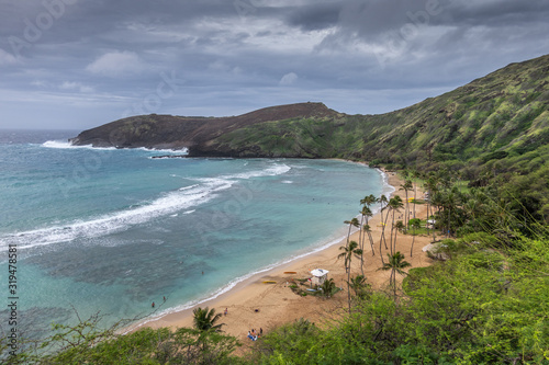 Oahu, Hawaii, USA. - January 11, 2020: Hanauma Bay Nature Preserve. Overview of Brown sandy beach with palmtrees, white surf on azure water, dark cliffs and rocks. All under heavy storm cloudscape.