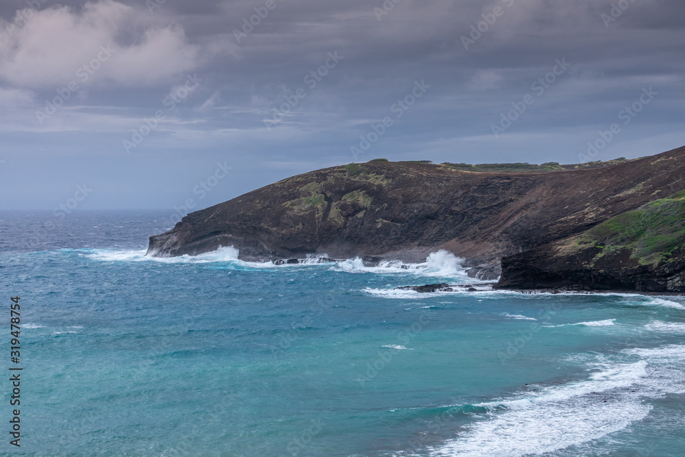 Oahu, Hawaii, USA. - January 11, 2020: Hanauma Bay Nature Preserve. White waves of azure water run along cliffs with face of baboon under heavy rain cloudscape. Oahu, Hawaii, USA. - January 11, 2020: Hanauma Bay Nature Preserve. White waves of azure water run along cliffs with face of baboon under heavy rain cloudscape.