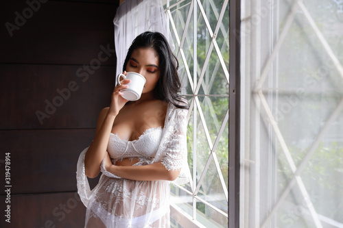 Portrait of attractive and sexy asian woman in white lingerie sleepwear standing beside window and drinking coffee in the morning with relaxation. Young cute girl long hair standing in bedroom © feeling lucky