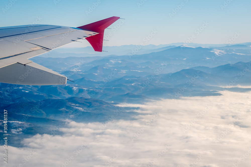 Above the clouds. Wing aircraft in altitude during flight, alternating mountain ranges partially covered with thick white clouds, clear blue sky in the bckground. Flight over the Balkans. Above the clouds. Wing aircraft in altitude during flight, alternating mountain ranges partially covered with thick white clouds, clear blue sky in the bckground. Flight over the Balkans.
