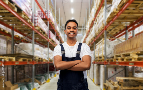 logistic business and people concept - happy smiling indian loader or worker with crossed arms over warehouse background