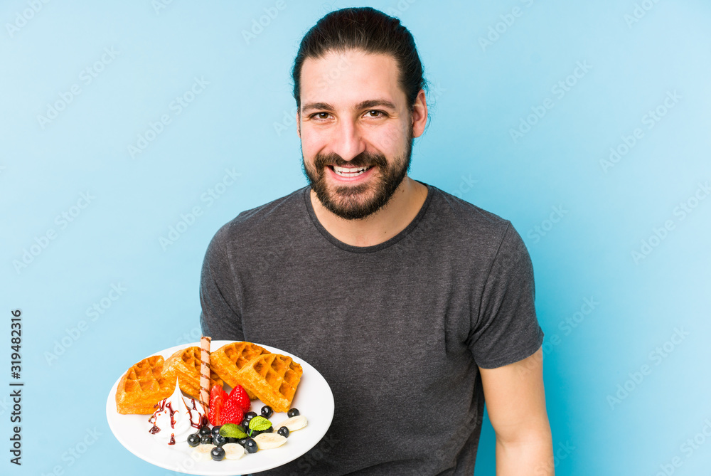 Young caucasian man eating a waffle dessert isolated laughing and having fun. Young caucasian man eating a waffle dessert isolated laughing and having fun.