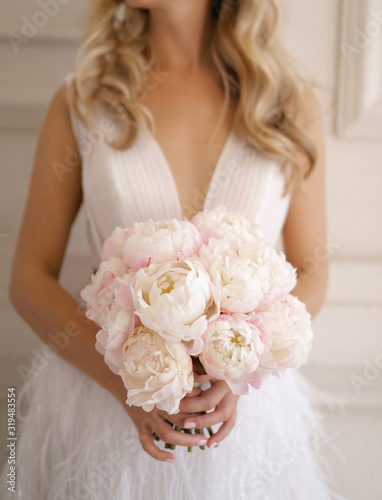 The bride in a chic white dress holds a bouquet of fresh blooming peonies in white and pink colors.