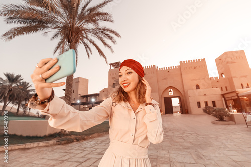 Photography Happy woman traveler wearing dress and turban taking selfie photos on her smartphone of an old Arab town or village in the middle of the desert