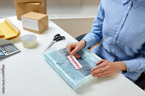 delivery, mail service, and shipment concept - close up of woman sticking fragile mark to parcel in protective bubble wrap at post office