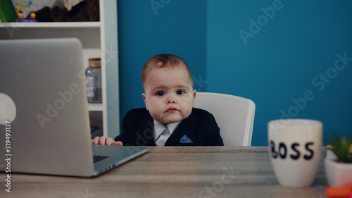 Cute baby boss working with laptop sitting on the chair at the office. Tapping his hands, enjoying the play, humor. Positive emotions, lovely child’s portrait. Close up view, selective focus