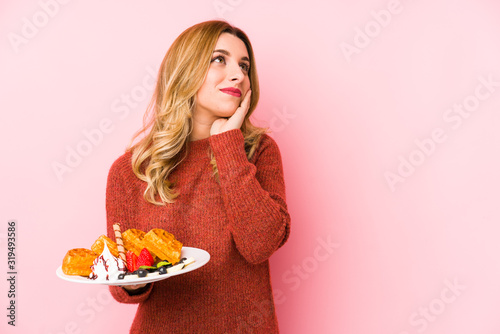 Young blonde woman eating a waffle dessert isolated looking sideways with doubtful and skeptical expression.