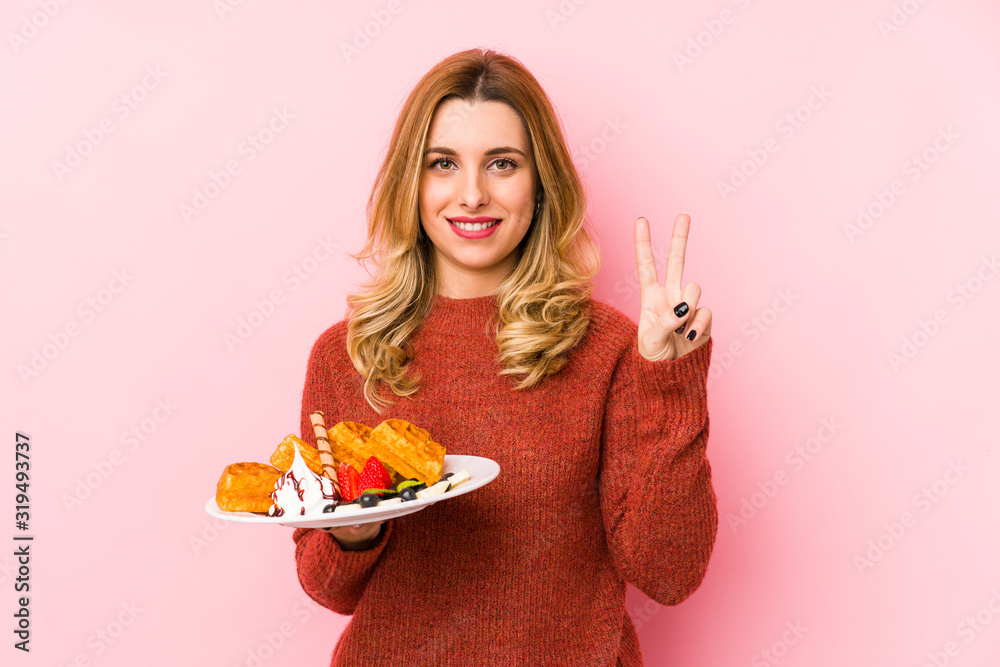 Young blonde woman eating a waffle dessert isolated showing number two with fingers. Young blonde woman eating a waffle dessert isolated showing number two with fingers.